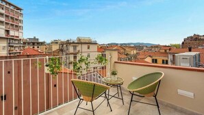 Un balcón con dos sillas de mimbre y una pequeña mesa, con vista a un paisaje urbano con edificios y una cadena montañosa distante.