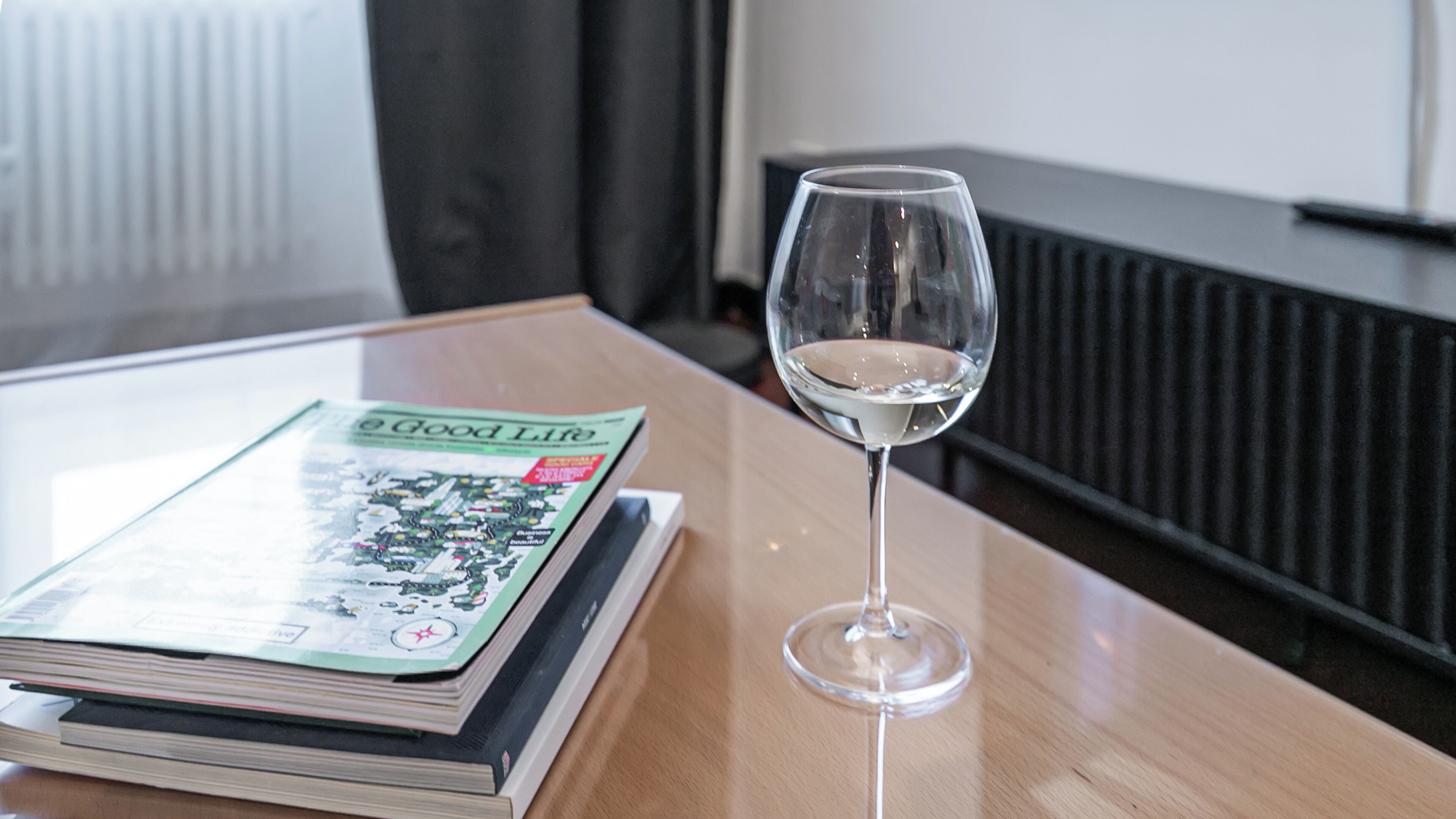 A glass of white wine on a wooden table with a magazine titled "Good Life" and a book.