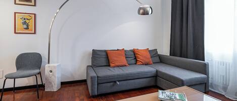 Un salon moderne avec un canapé d’angle gris, deux coussins orange, un fauteuil gris, une table d’appoint blanche sur laquelle reposent un magazine et un verre de vin, un lampadaire et deux tableaux encadrés accrochés au mur.
