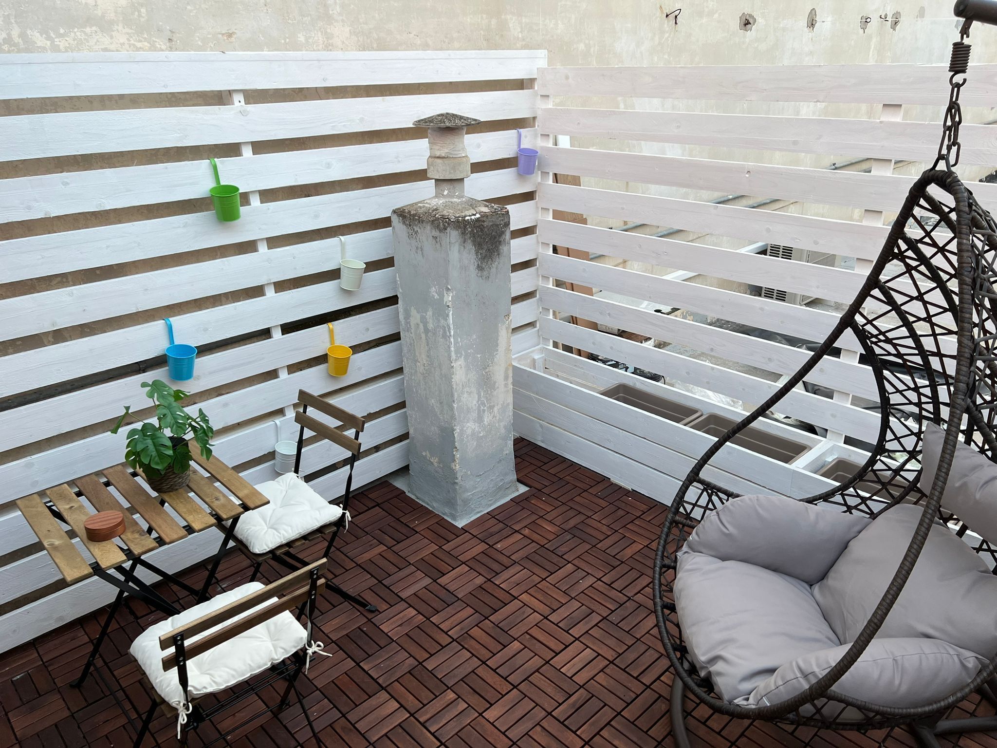 Un patio con una silla colgante, una mesita con una planta y un separador blanco con contenedores de almacenamiento.