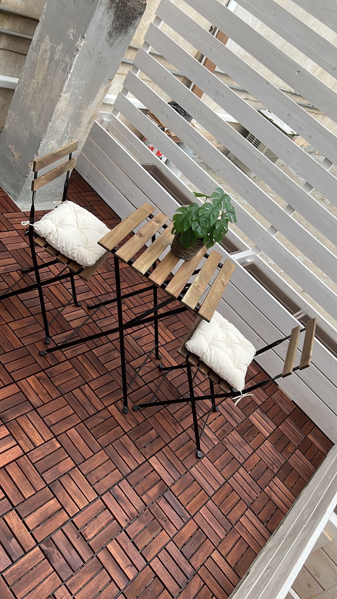 A wooden table with two cushioned chairs and a potted plant, set on a brick patio with a metal railing in the background.