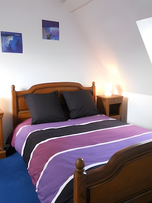 A bedroom with a wooden bed, two pillows, a bedside table with a lamp, and a wall-mounted artwork.