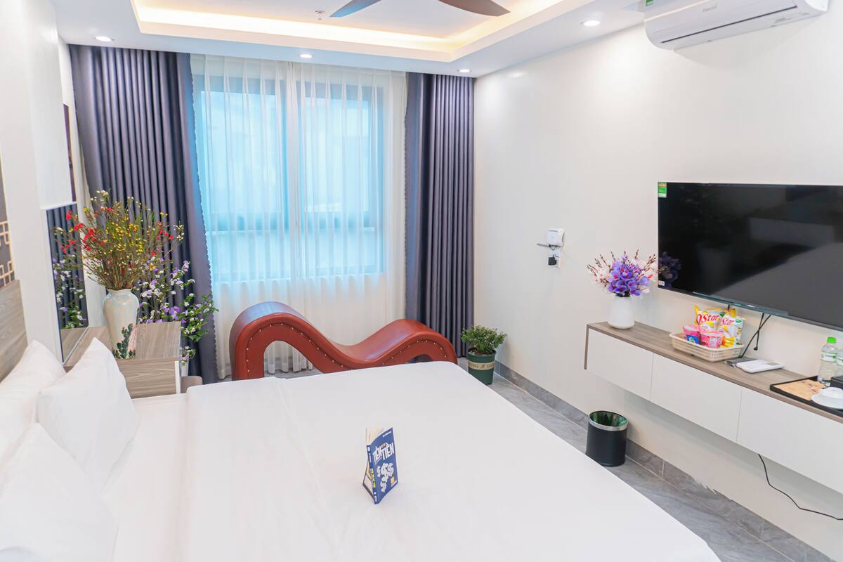 Una habitación de hotel moderna con una cama grande, televisor, escritorio con computadora y ventana con cortinas.