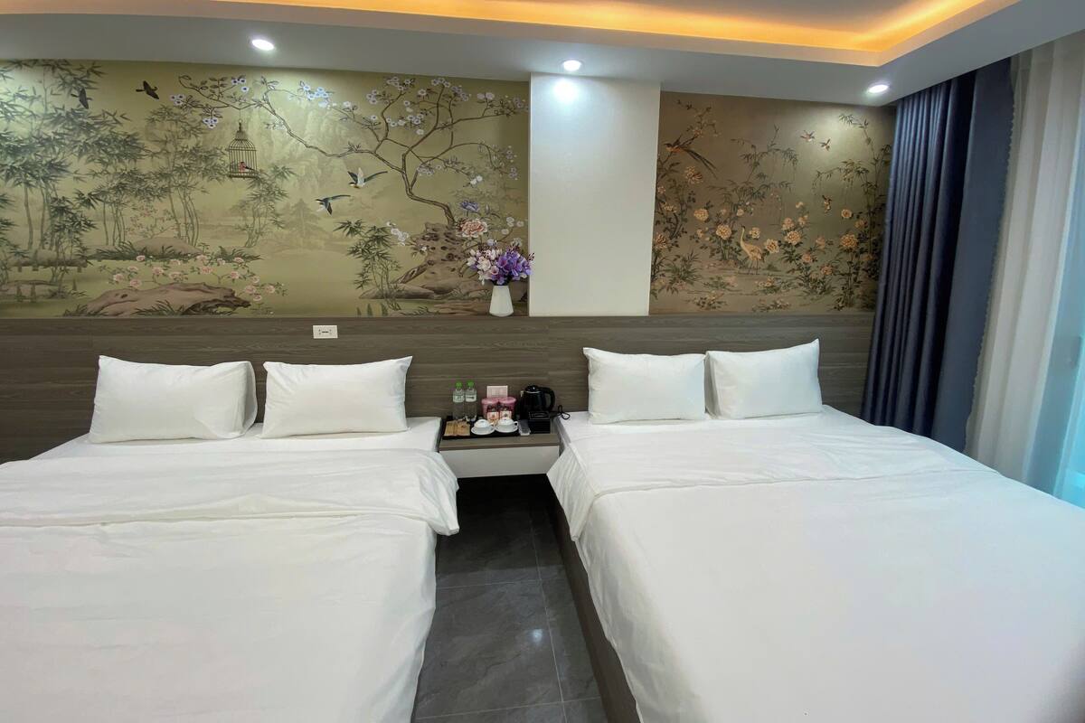 Habitación de hotel con dos camas, una mesita de noche con artículos de aseo, un mural decorativo y una ventana con cortinas.