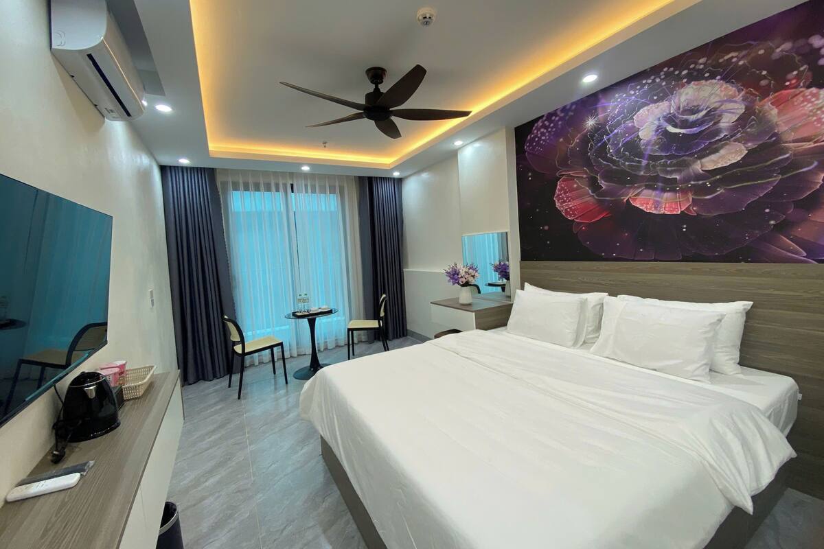 Habitación de hotel con cama, un escritorio con silla, una pequeña mesa con sillas, un ventilador de techo y un mural grande en la pared.