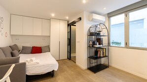 Une chambre moderne avec un lit, un canapé gris, une bibliothèque et une fenêtre avec des stores.