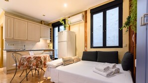 Une chambre d’hôtel compacte comprenant un lit, une petite table recouverte d’une nappe à motifs floraux, un coin cuisine et une fenêtre donnant sur un paysage agréable.