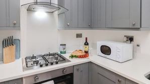 Een keuken met grijze kastjes, een gasfornuis, een magnetron en een klein aanrecht met een brood, tomaten en een fles wijn.