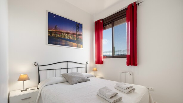 Un lit bien fait, avec du linge de lit et des serviettes blanches, une table de chevet avec une lampe, un tableau encadré représentant un pont, et une fenêtre avec des rideaux rouges.