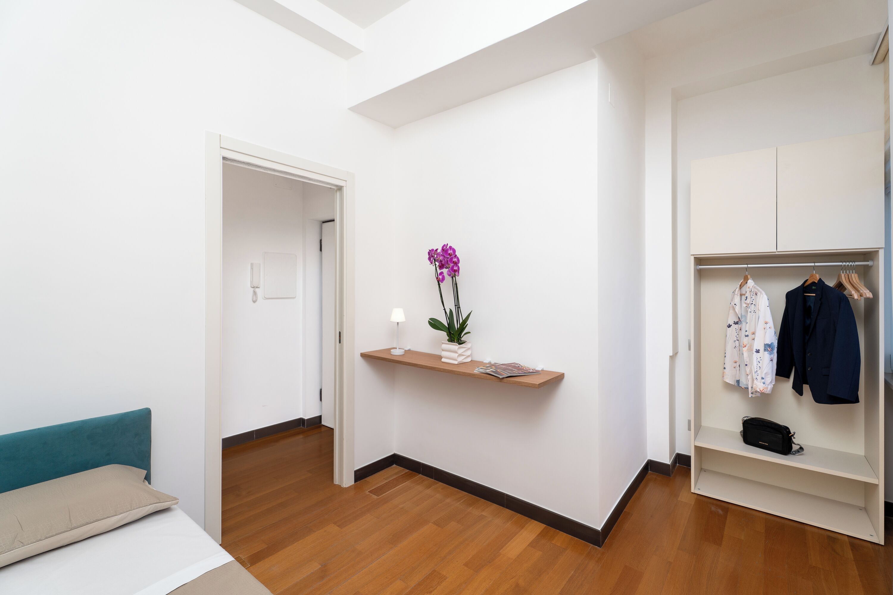 A hotel room with a bed, a wooden floor, a white wardrobe, a shelf with a plant, and a door leading to another room.