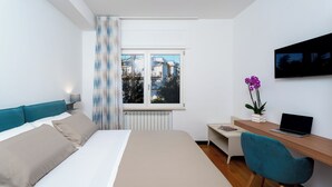 A modern hotel room with a bed, a desk with a laptop, a TV, and a window with a view of buildings. - YourHome - Lady Lucy Holiday Home (Meta)