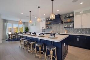 Uma cozinha moderna com uma ilha grande, banquetas de bar, luminárias pendentes e prateleiras decorativas.