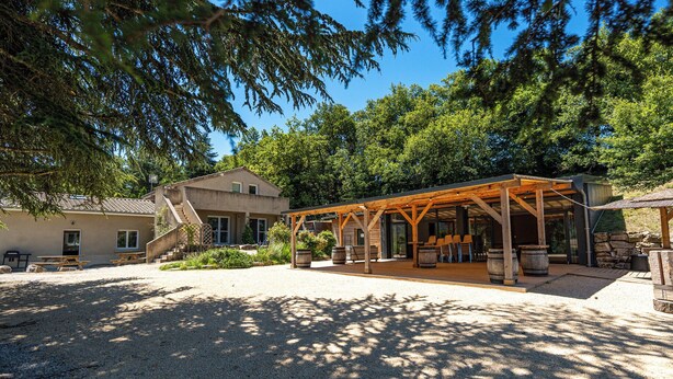 Un espace extérieur aménagé dans un style rustique, avec des tables et des bancs en bois, entouré d’arbres et bordé d’un bâtiment doté d’un mur en pierre.