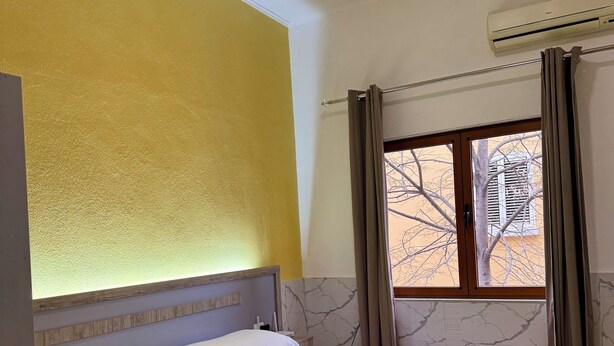 A bedroom with a bed, a window with curtains, an air conditioning unit, and a view of trees outside. - Tirana Central Apartment - Free Parking - Fit Five (Tiranë)