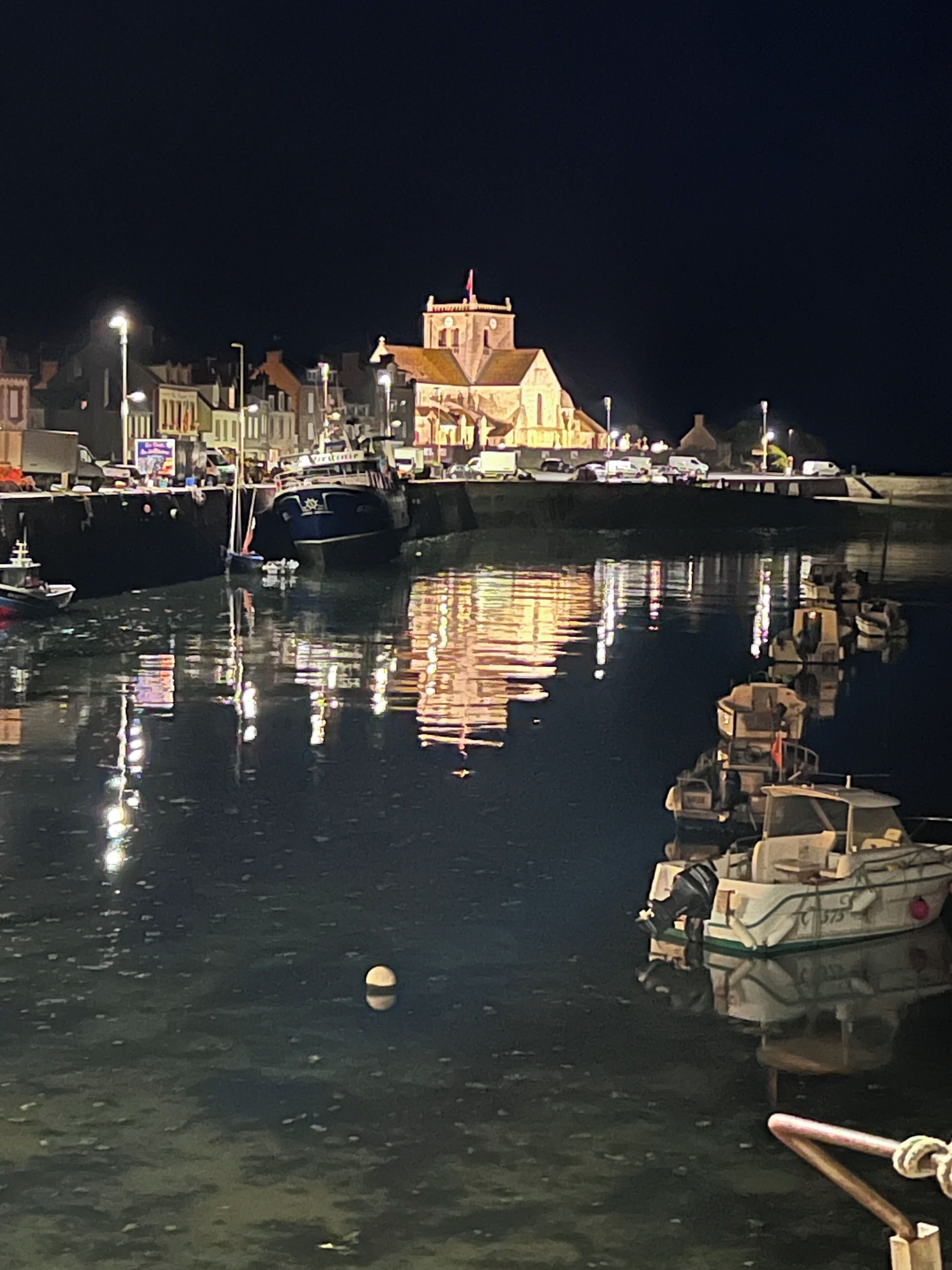 Un puerto nocturno con barcos amarrados, una iglesia con campanario y edificios a lo largo del muelle.