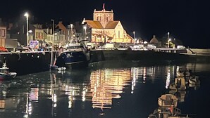 Um porto noturno com barcos atracados, uma igreja com campanário e edifícios ao longo da orla.