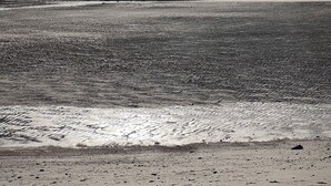 Uma praia com mar calmo, uma construção ao longe e céu nublado.
