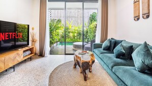 A modern living room with a TV, sofa, and a view of a garden through glass doors. - Ocean Breeze Berawa Villa (Canggu)