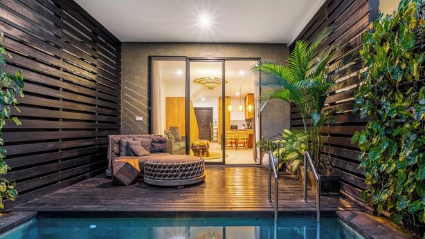 A modern outdoor pool area with a wooden deck, a round seating area with cushions, and a glass door leading to a kitchen and dining area. - Ocean Breeze Berawa Villa (Canggu)