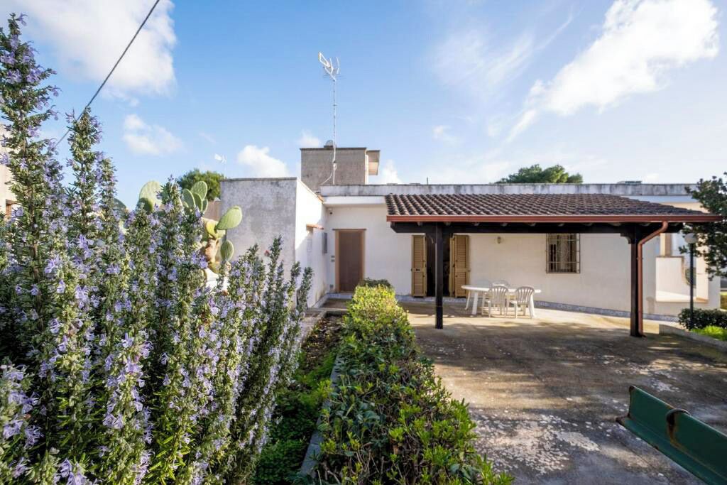 A building with a tiled roof, a covered patio with chairs, and a garden with purple flowers.