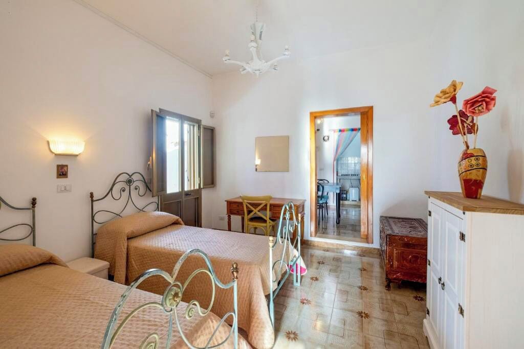 A bedroom with two beds, a desk, and a chair. There is a mirror on the wall and a vase with flowers on a cabinet.