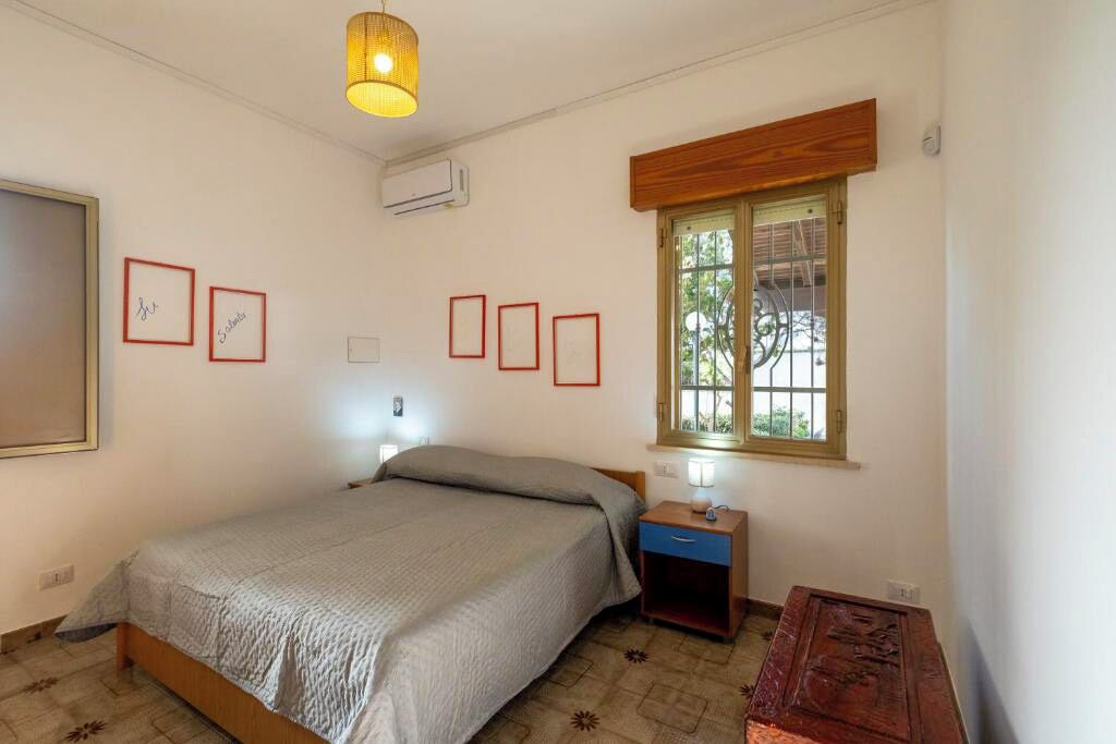 A bedroom with a bed, bedside table, lamp, window, and wall-mounted air conditioner.