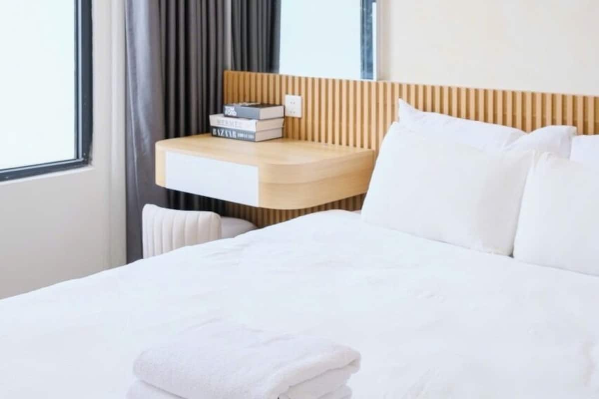 A hotel room with a bed, a bedside table with books, a mirror, and a window with curtains.