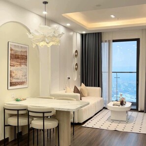 A modern living room with a sofa, a dining area with a table and chairs, a large window with a city view, and a decorative light fixture. - Pi & Pu Apartment - Hoang Huy Commerce (Hai Phong)