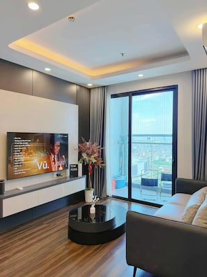 A modern living room with a flat-screen TV, a coffee table, a sofa, and a view of the city through large windows. - Pi & Pu Apartment - Hoang Huy Commerce (Hai Phong)