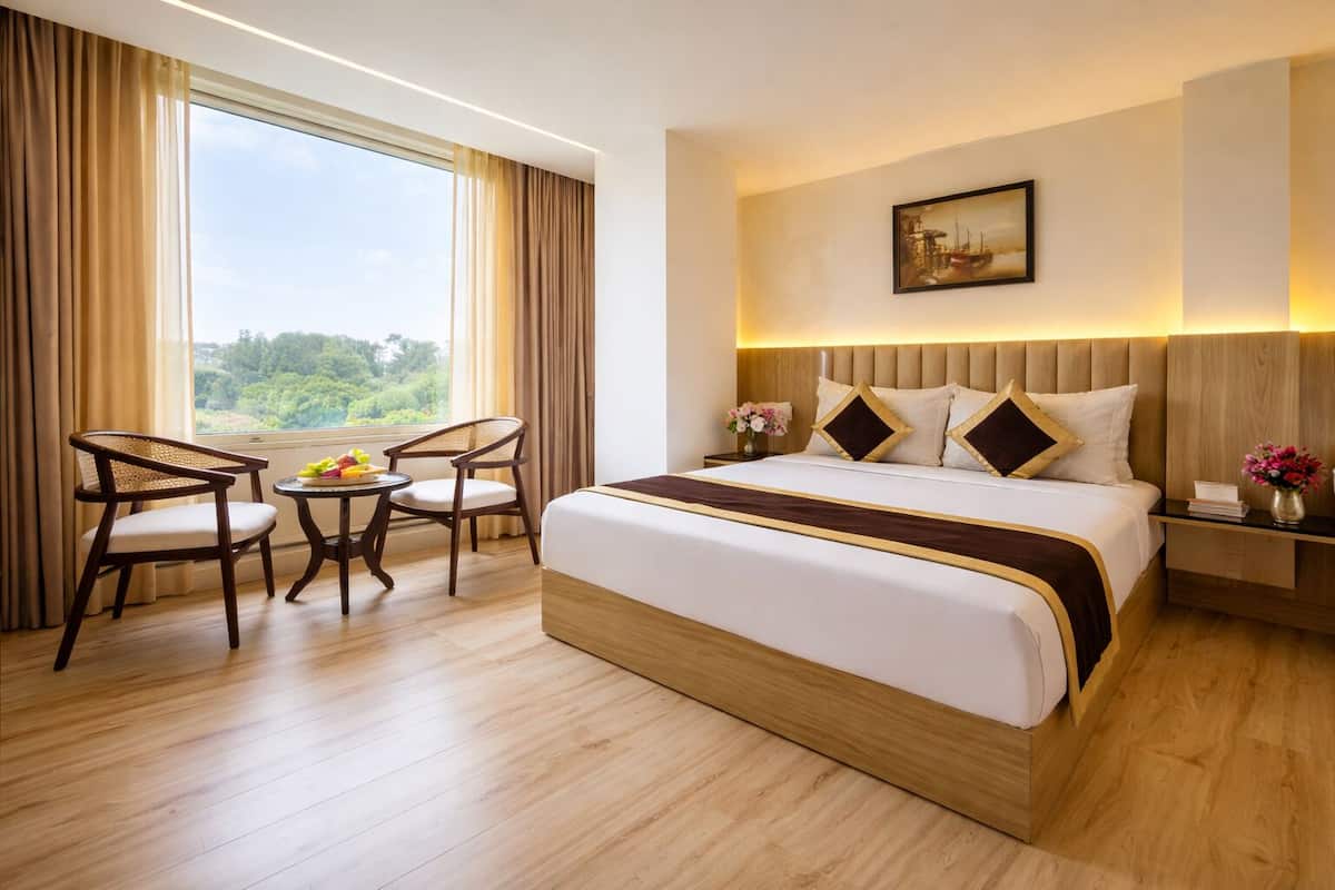 Habitación de hotel con una cama grande, dos sillas, una mesita con fruta, una mesita de noche con un jarrón y un cuadro en la pared.