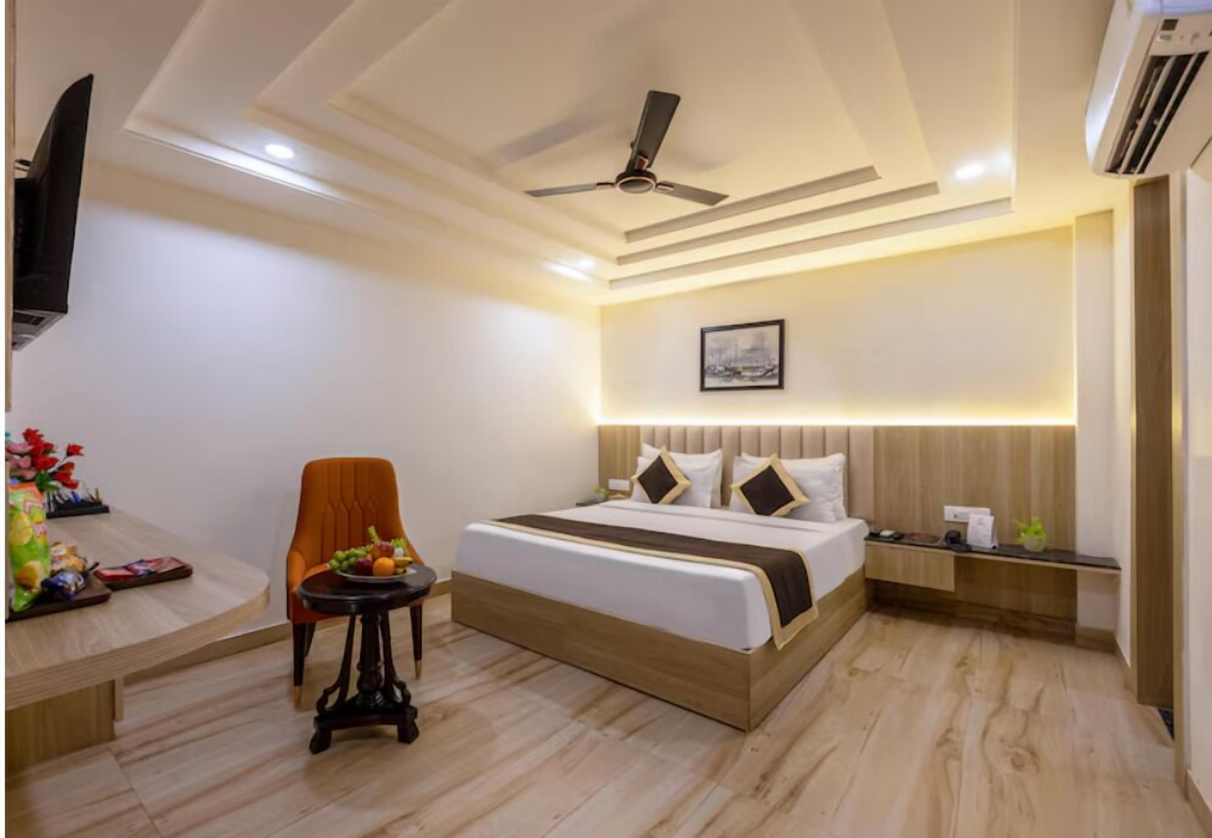 A modern hotel room with a bed, a chair, a small table with fruits, a ceiling fan, and an air conditioning unit.