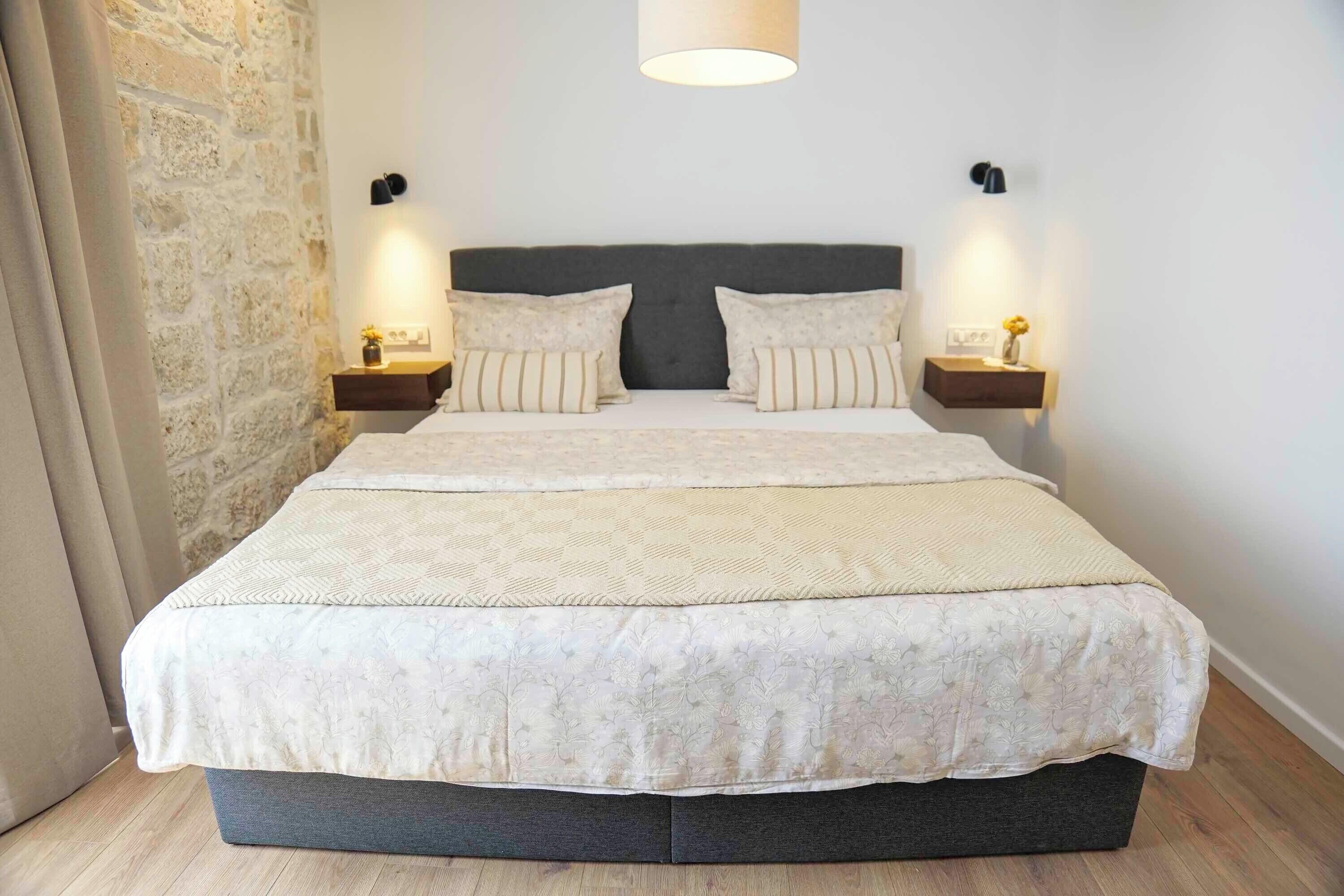 A bedroom with a large bed, two bedside tables with lamps, a stone wall, and a wooden floor.
