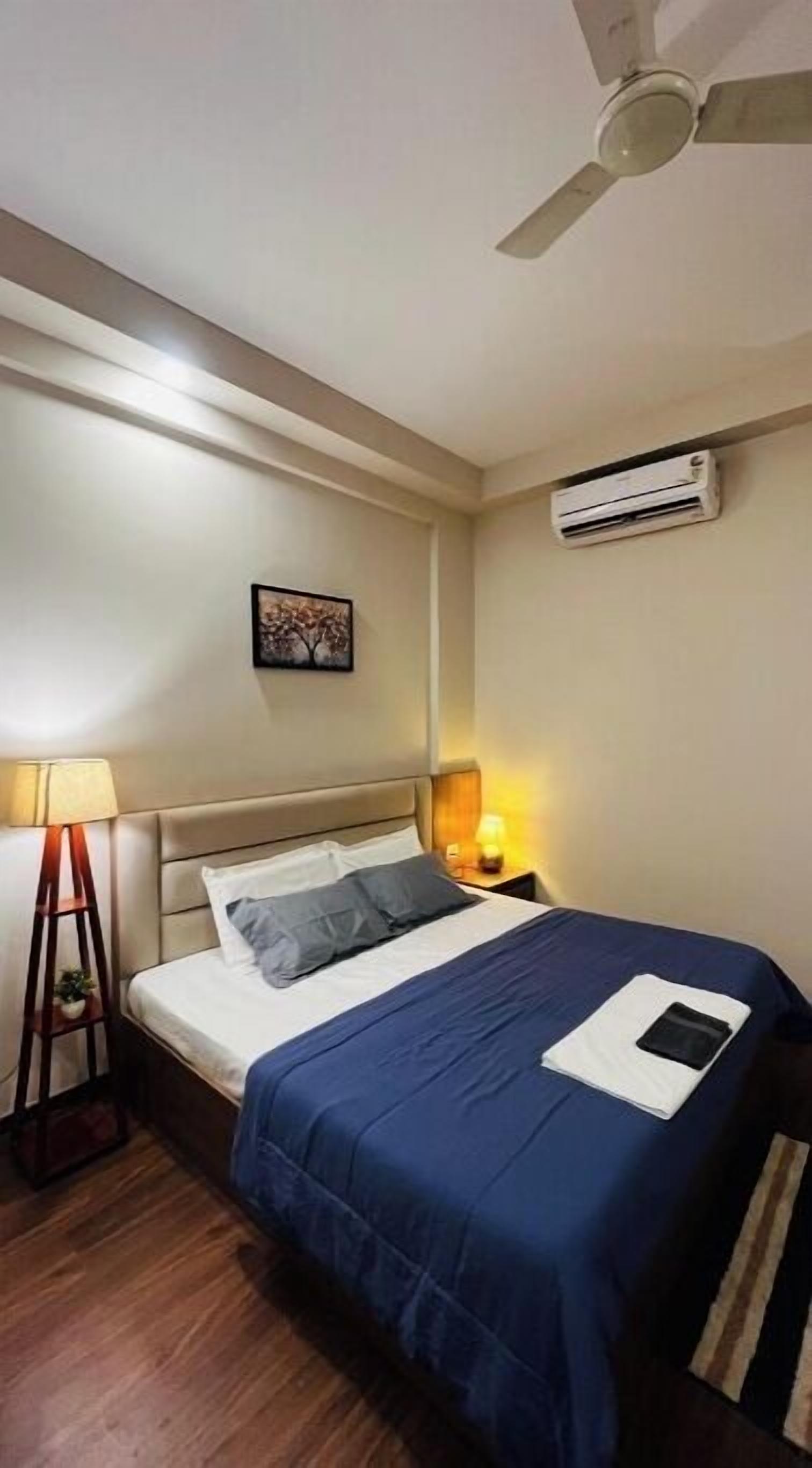 Una habitación de hotel con una cama, ventilador de techo, equipo de aire acondicionado, mesita de noche con lámpara y un cuadro en la pared.