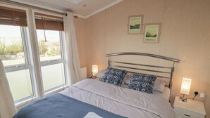 A neatly made bed with a blue and white duvet, two pillows with a geometric pattern, and two folded towels on a blue bed runner. There are two bedside tables with lamps, a window with curtains, and two framed pictures on the wall. - Rhosgranog Caravan (Haverfordwest)