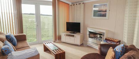 Ein geräumiges Wohnzimmer mit Fernseher, Kamin und Blick auf ein grünes Feld durch große Fenster.