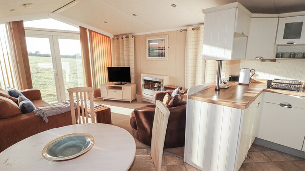 A modern living room with a sofa, TV, and fireplace. There is a dining area with a table and chairs. The kitchen has white cabinets and a coffee maker. - Rhosgranog Caravan (Haverfordwest)