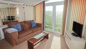 A modern living room with a brown sofa, a wooden coffee table, a TV, and large windows offering a view of greenery. - Rhosgranog Caravan (Haverfordwest)