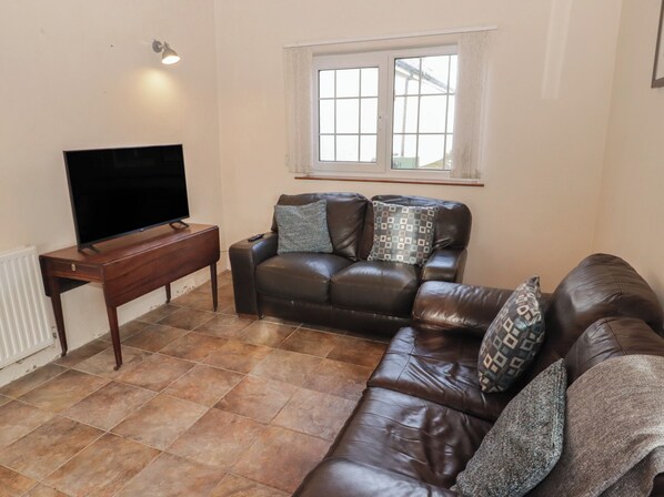A living room with a brown leather sofa, a wooden coffee table, a flat-screen TV, and a window with white curtains. - Farm Cottage (Haverfordwest)