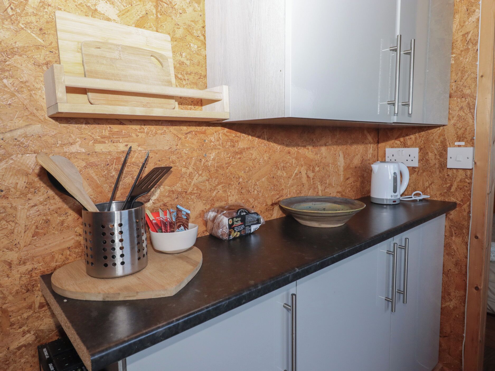 A kitchen with a wooden cutting board, a metal utensil holder, a bowl, a bag of snacks, a coffee maker, and a cabinet.