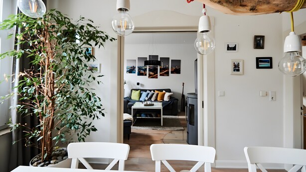 A modern living room with a sofa, dining table, and chairs, a large plant, and hanging light fixtures. - Unique Carpenter’s Apartment with Sea View, Balcony, and Wi-Fi (Oldenburg)