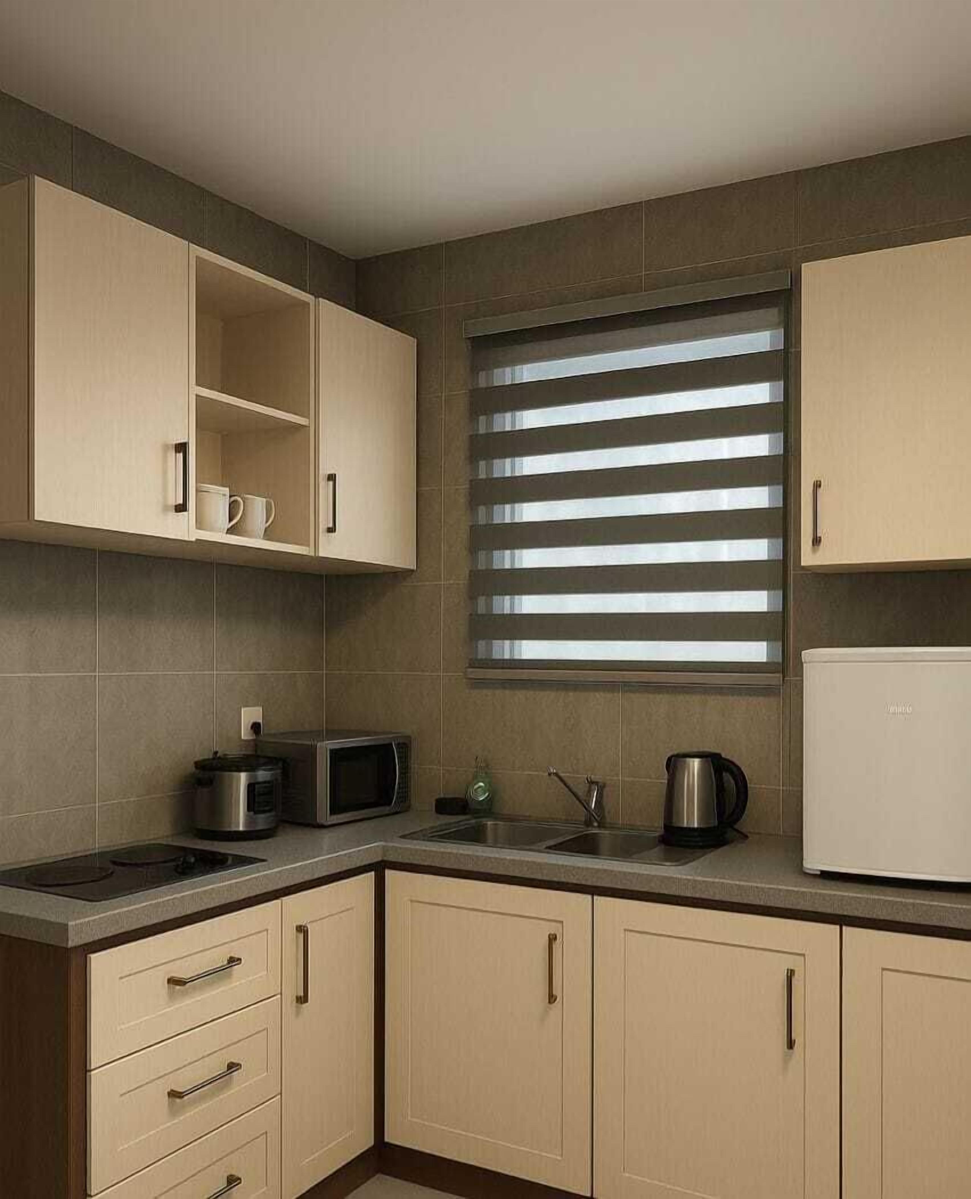 A kitchen with a microwave, kettle, and a small refrigerator. There are cabinets and a window with blinds.