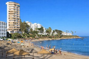 Ein Strand mit einem hohen Hotel, Palmen und Sonnenschirmen.