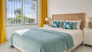 A bedroom with a bed, two nightstands, a window with yellow curtains, and a view of greenery outside. - Sunny Garden Apartment Miraflores (Mijas Costa)