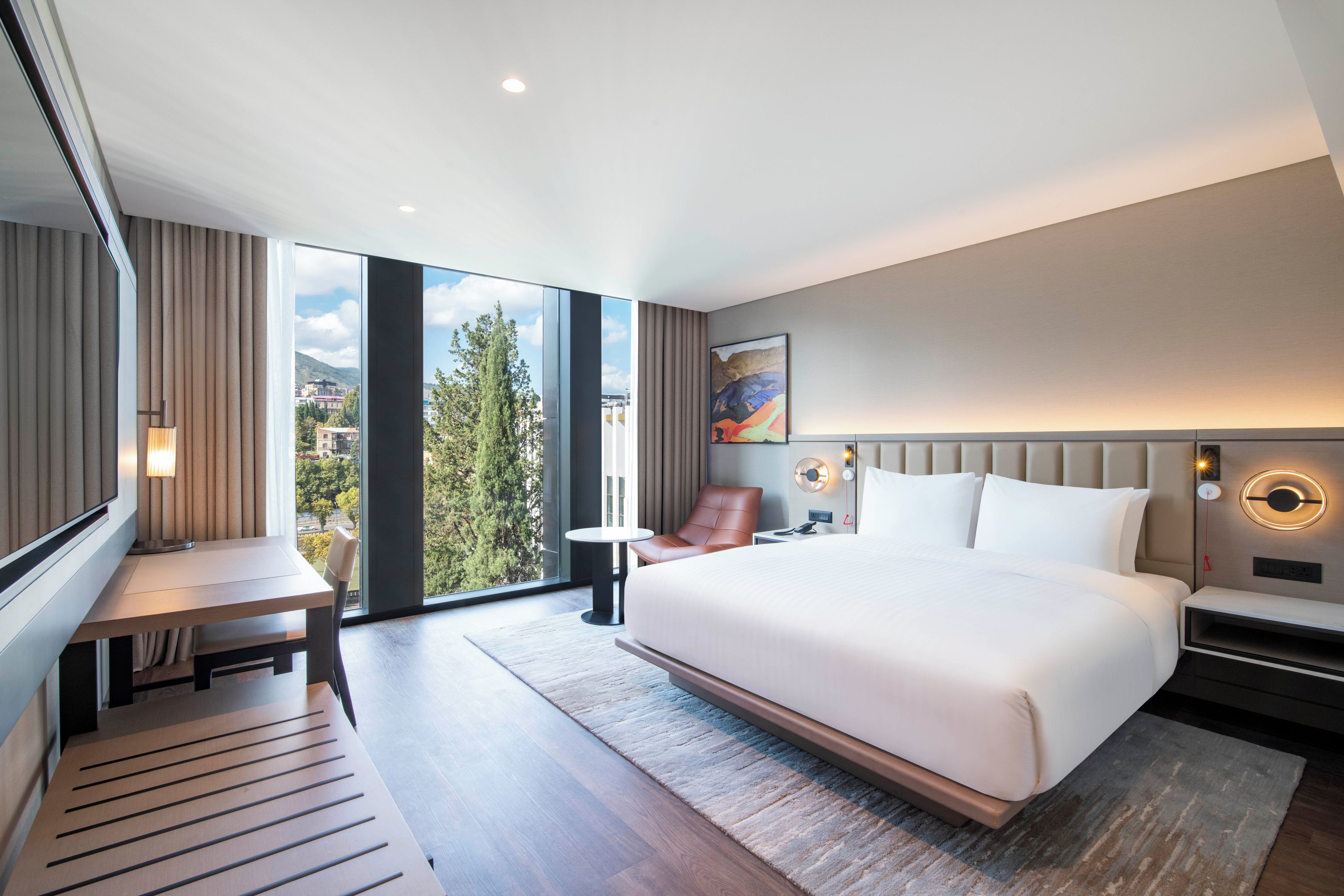 Quarto de hotel moderno com uma cama grande, uma escrivaninha, uma cadeira e vista para o exterior.