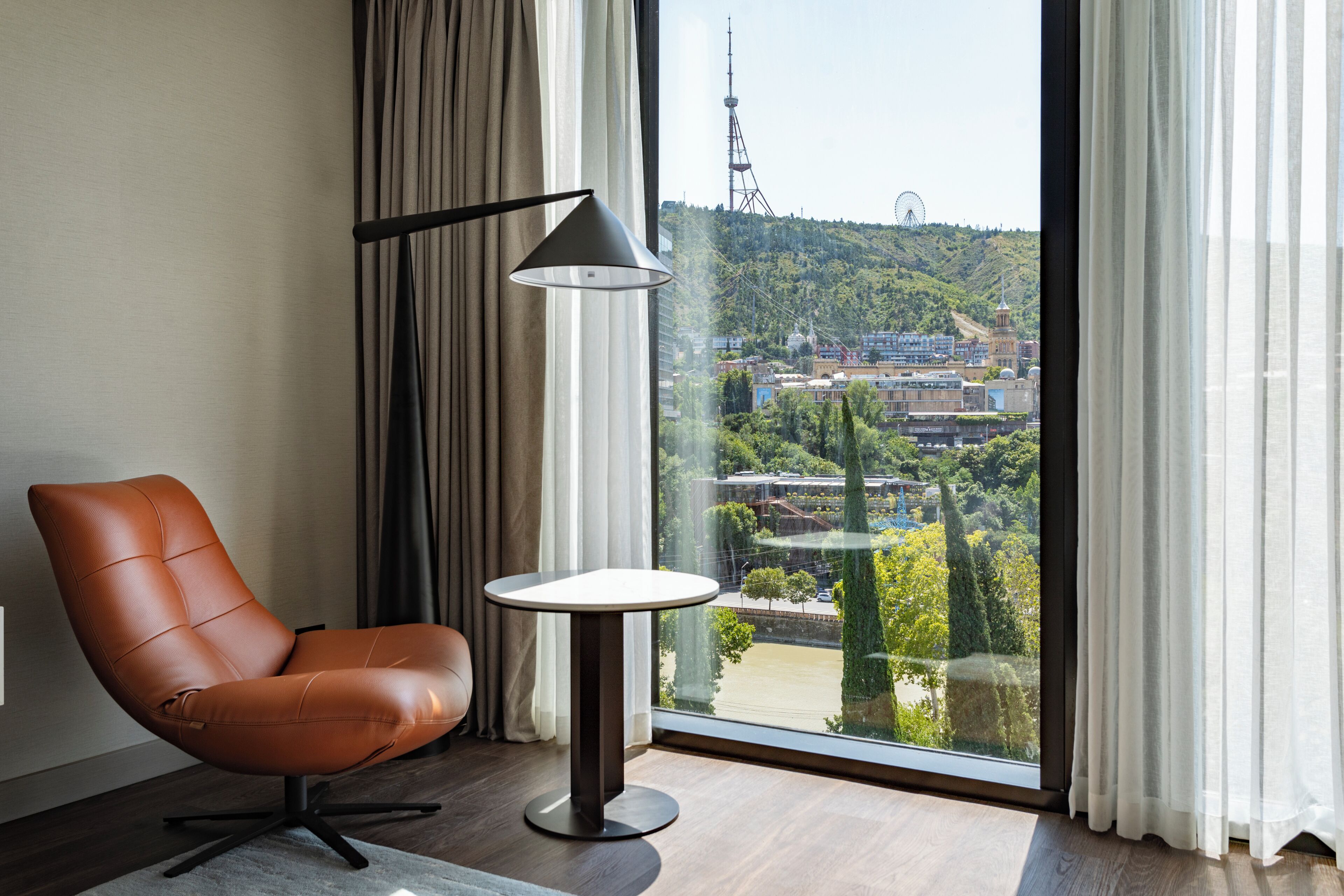 A modern room with a leather chair, a small table, and a view of a cityscape through large windows.