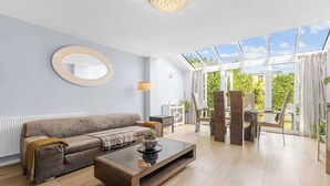 A modern living room with a sofa, coffee table, and a dining area with a table and chairs. A large window provides a view of greenery outside. - 3-bed Greenhithe Retreat Garden Near Bluewater (Greenhithe)