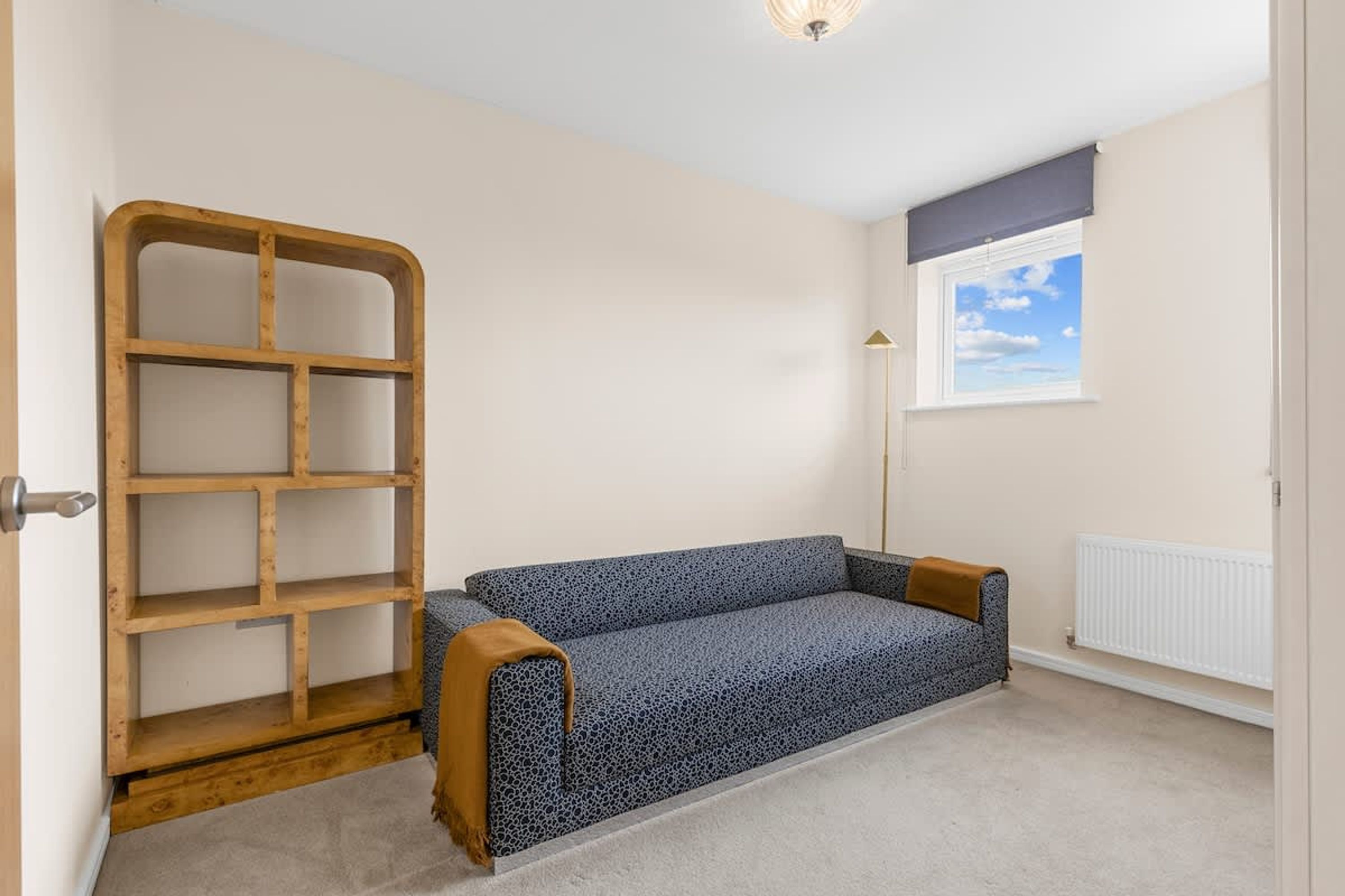A room with a blue patterned sofa, a wooden shelf unit, a radiator, and a window with a view of the sky.