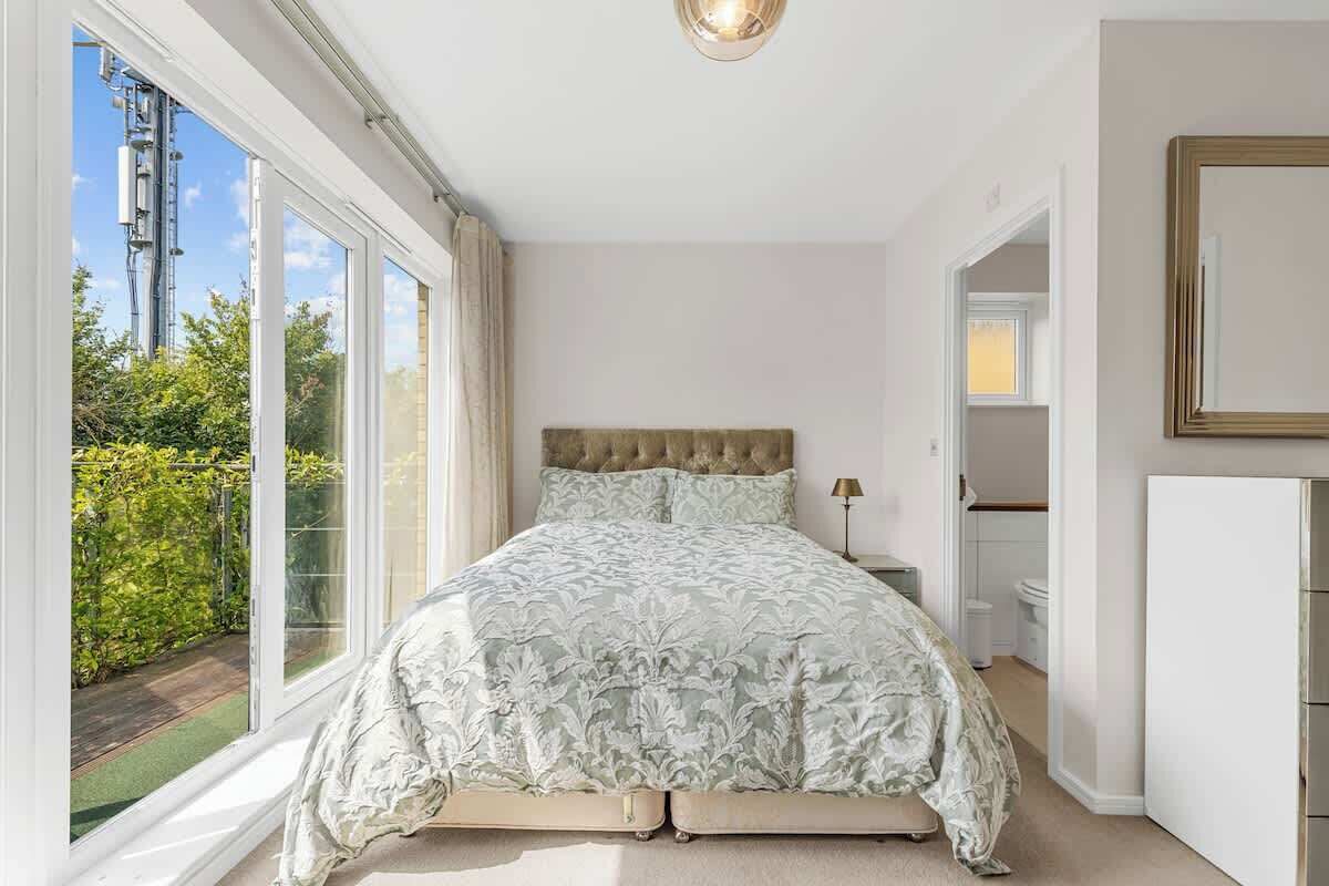 A bedroom with a large window, a bed with a tufted headboard, and a bedside table with a lamp.
