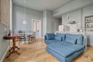 A modern living room with a blue sofa, wooden dining table, and chairs. A kitchen area with white cabinets and a countertop is visible in the background. - w Bright 2BR Near Arco Della Pace (Milan)