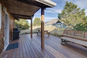Una terraza de madera con mesas de picnic y un banco, con vistas a un área verde y una casa.